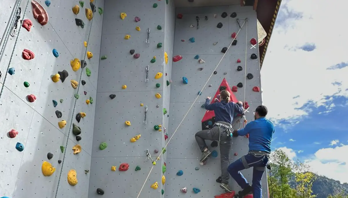 Zwei Personen klettern an einer bunten künstlichen Kletterwand im Freien, eine dritte Person sichert. | © DAV Dortmund