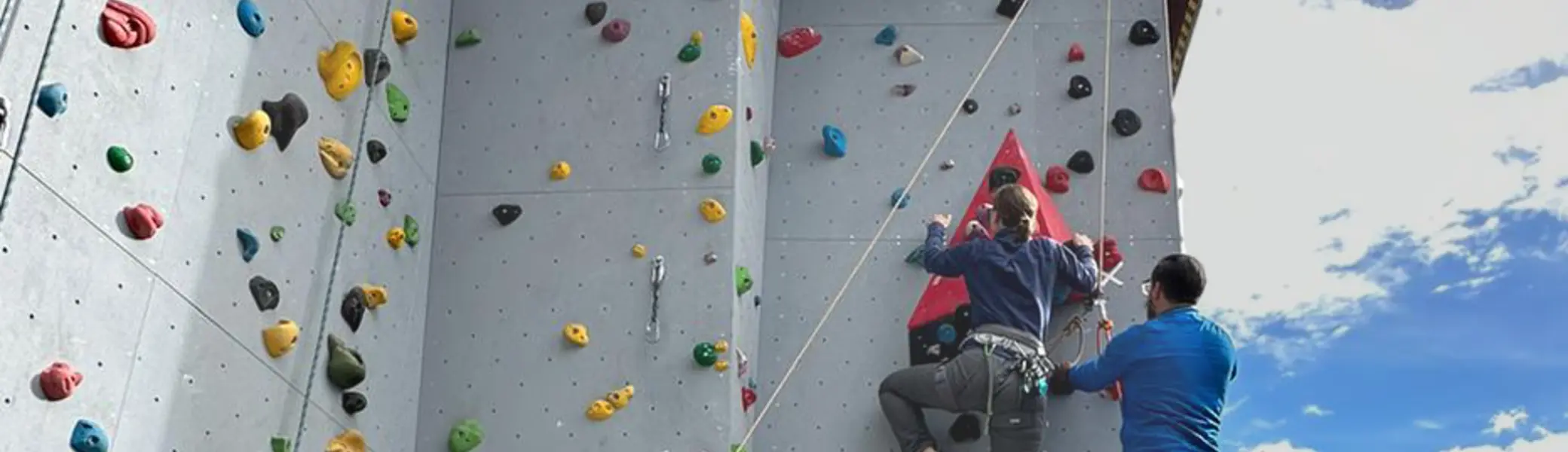 Zwei Personen klettern an einer bunten künstlichen Kletterwand im Freien, eine dritte Person sichert. | © DAV Dortmund