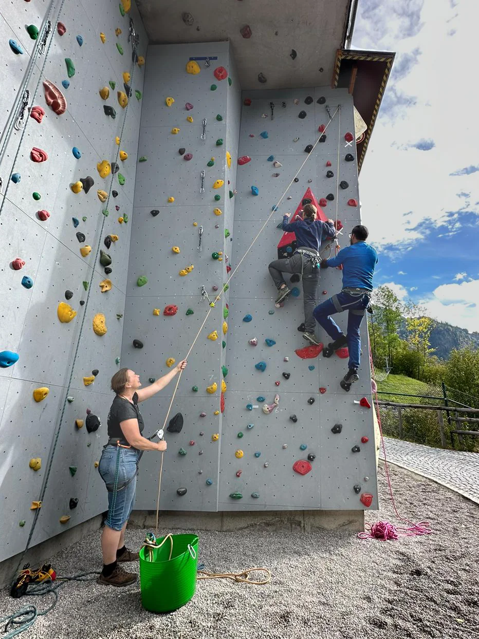 Zwei Personen klettern an einer bunten künstlichen Kletterwand im Freien, eine dritte Person sichert. | © DAV Dortmund