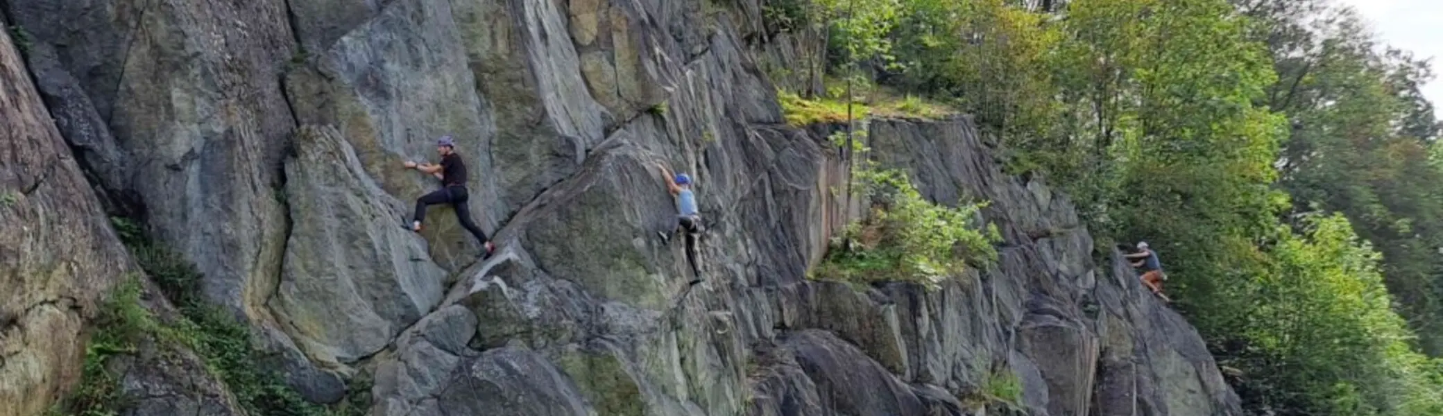 Personen klettern an einer steilen, dunklen Felswand direkt neben einem Wanderweg im Grünen. | © JDAV