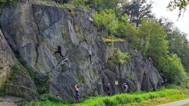 Personen klettern an einer steilen, dunklen Felswand direkt neben einem Wanderweg im Grünen. | © JDAV