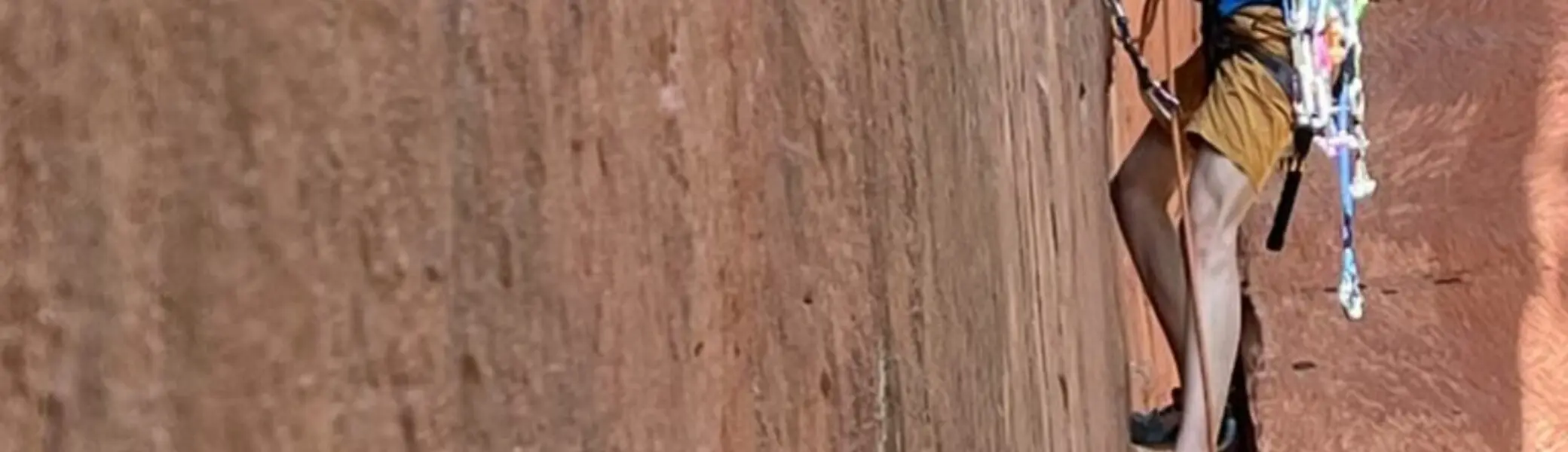 Kind mit Klettergurt und Helm klettert eine steile, rote Felswand im Freien hinauf. | © JDAV