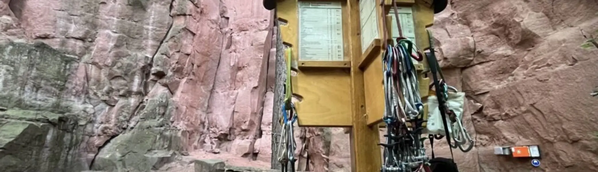 Holzständer mit zahlreichen Kletterausrüstungen vor einer hohen roten Felswand in einem Klettergebiet. | © JDAV