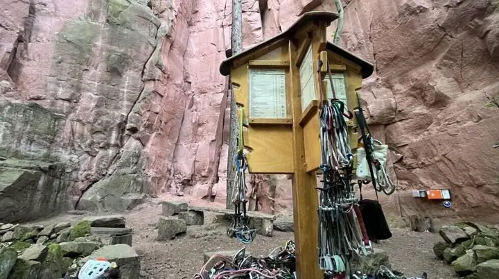 Holzständer mit zahlreichen Kletterausrüstungen vor einer hohen roten Felswand in einem Klettergebiet. | © JDAV