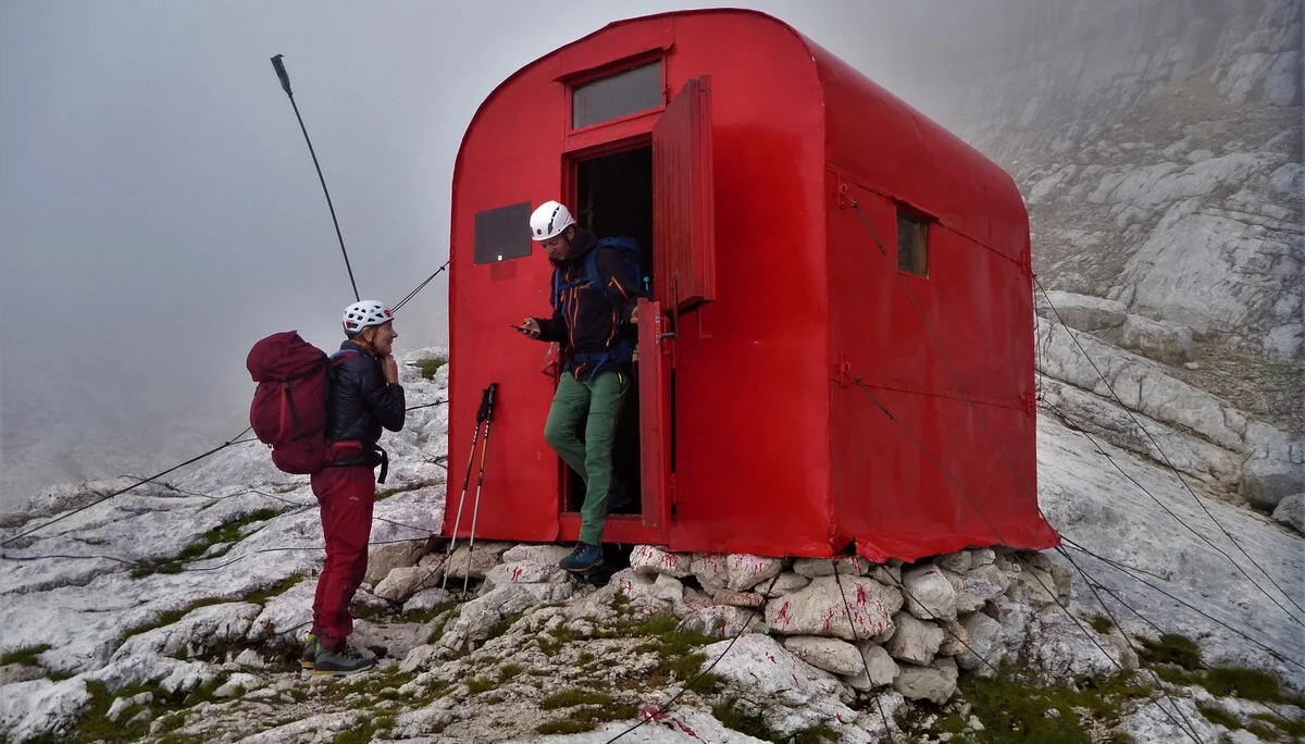 Zwei Personen stehen mit Rucksäcken vor einem roten Biwak auf felsigem Gelände bei Nebel. | © DAV Dortmund