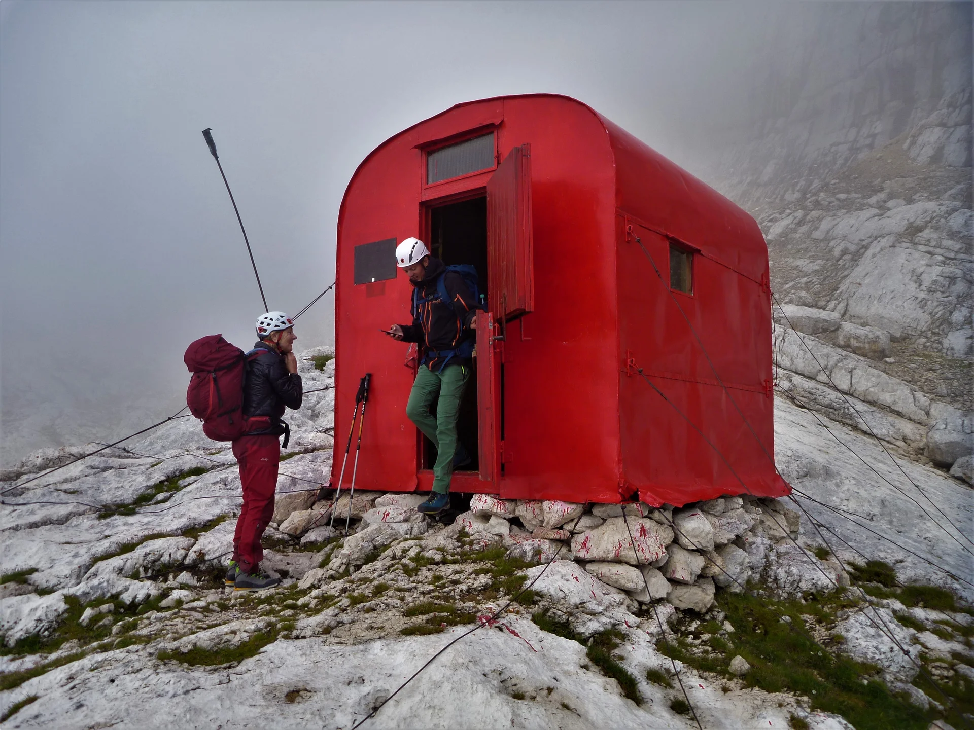 Zwei Personen stehen mit Rucksäcken vor einem roten Biwak auf felsigem Gelände bei Nebel. | © DAV Dortmund