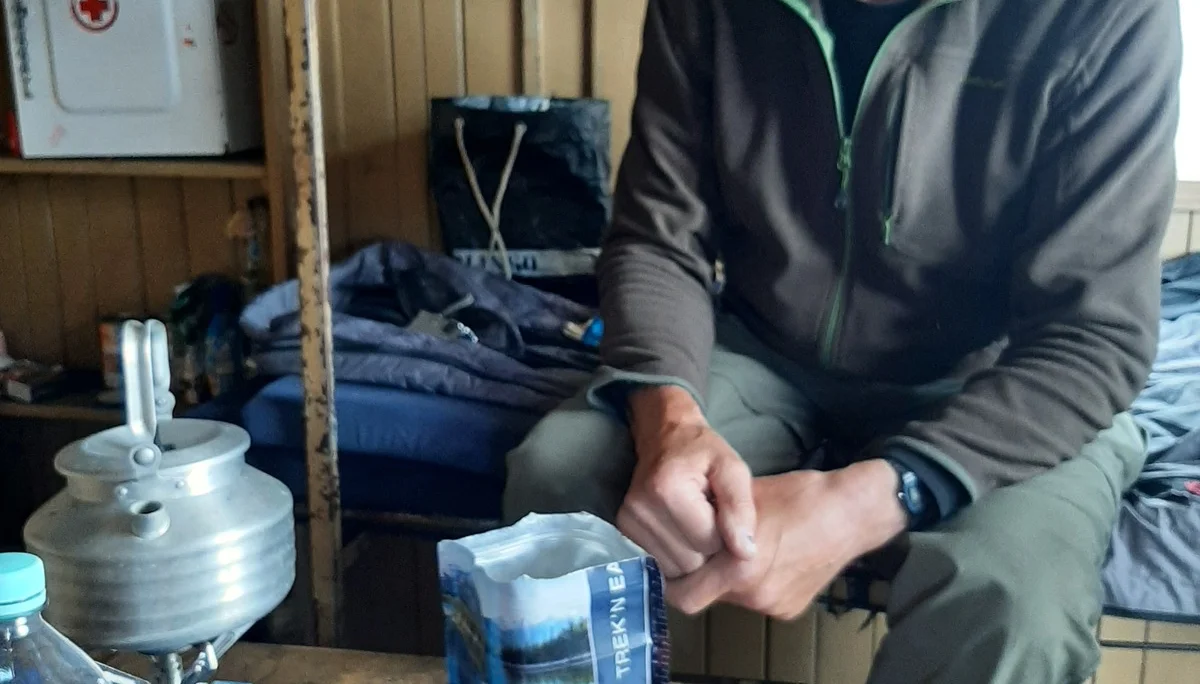 Eine Person sitzt an einem Tisch in einer einfachen Holzhütte mit Trekkingnahrung und Kocher. | © DAV Dortmund