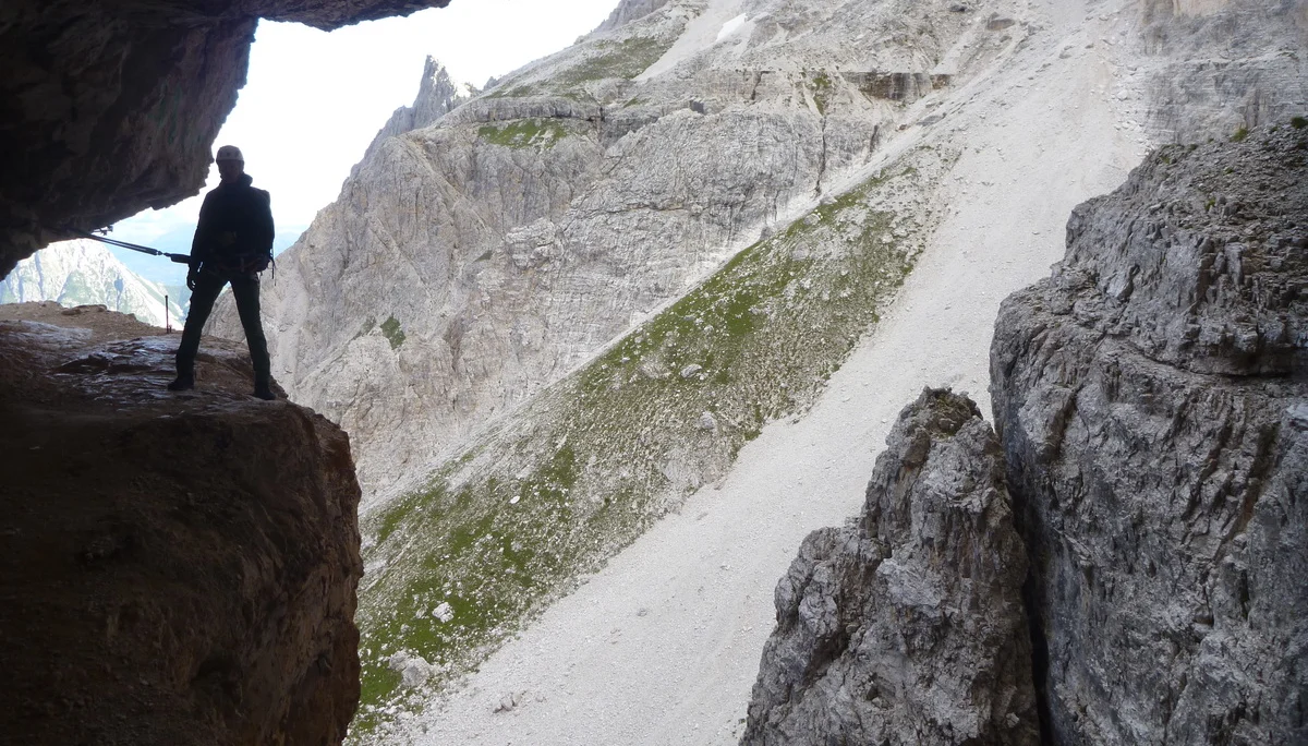 Eine Person steht am Eingang einer Felsgrotte mit Blick auf eine steile Geröllflanke in den Dolomiten. | © DAV Dortmund