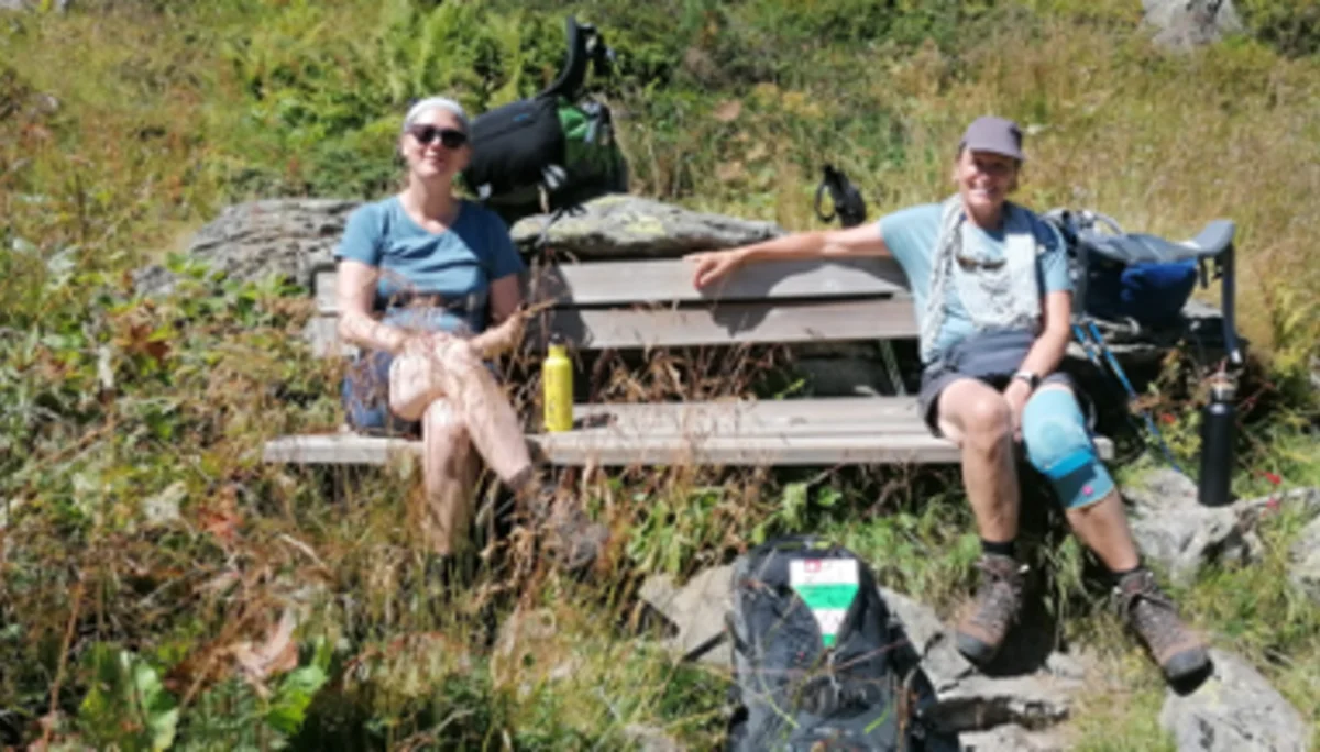 Zwei Personen sitzen auf einer Holzbank im Grünen, genießen eine Pause mit Rucksack und Getränk. | © DAV Dortmund