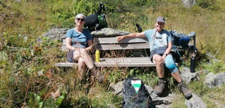 Zwei Personen sitzen auf einer Holzbank im Grünen, genießen eine Pause mit Rucksack und Getränk. | © DAV Dortmund