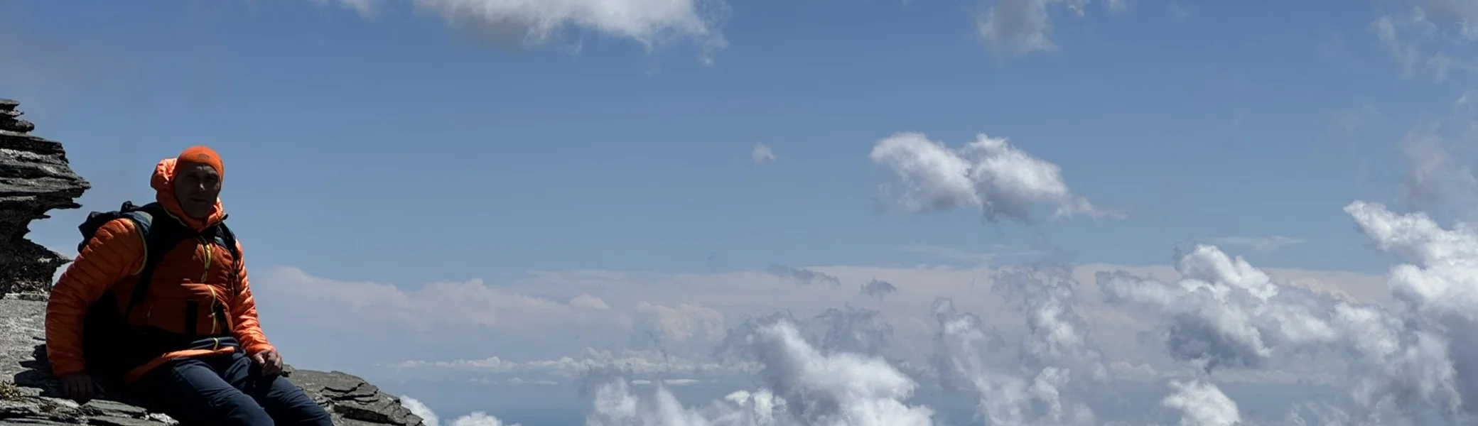 Eine Person in orangefarbener Jacke sitzt auf einem schmalen Felsvorsprung hoch über den Bergen, mit Wolken und blauem Himmel im Hintergrund. | © DAV Dortmund