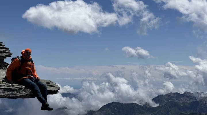 Eine Person in orangefarbener Jacke sitzt auf einem schmalen Felsvorsprung hoch über den Bergen, mit Wolken und blauem Himmel im Hintergrund. | © DAV Dortmund