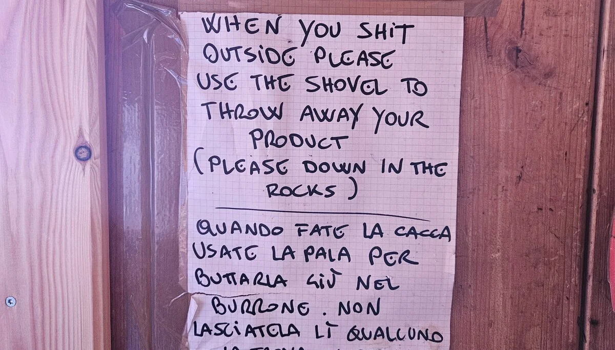 Handgeschriebener Zettel an einer Holzwand, der Besucher auffordert, beim Toilettengang draußen eine Schaufel zu benutzen und die Hinterlassenschaft in den Felsen zu entsorgen. Text auf Englisch und Italienisch. | © DAV Dortmund