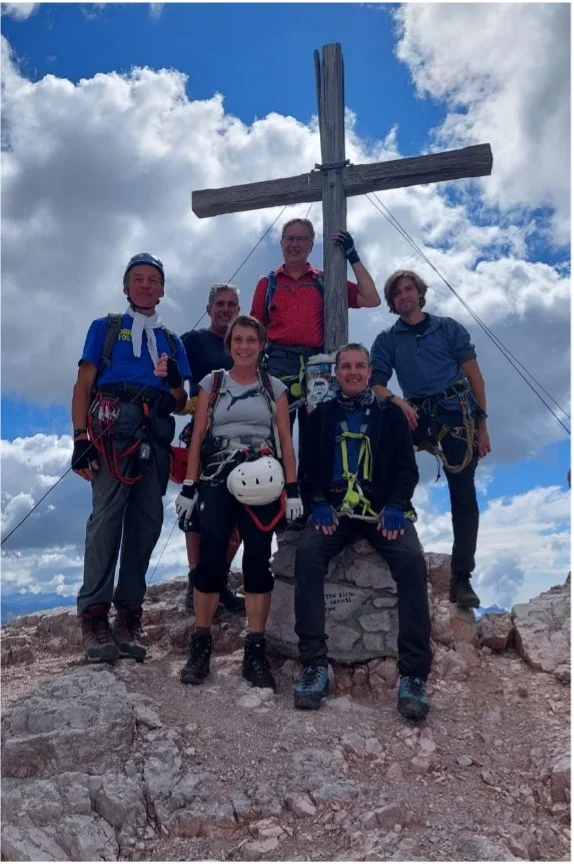 Sechs Personen stehen in Kletterausrüstung lachend am Gipfelkreuz unter wolkigem Himmel. | © DAV Dortmund