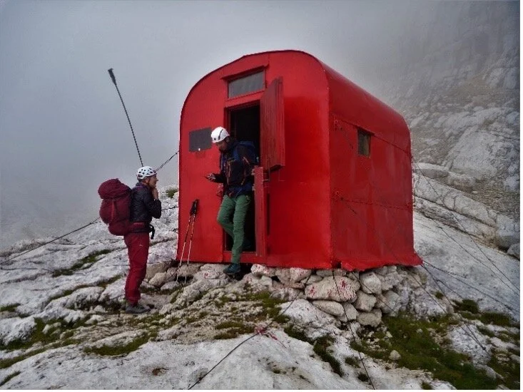Zwei Personen in Kletterausrüstung stehen vor einem kleinen roten Biwak in nebliger Umgebung. | © DAV Dortmund