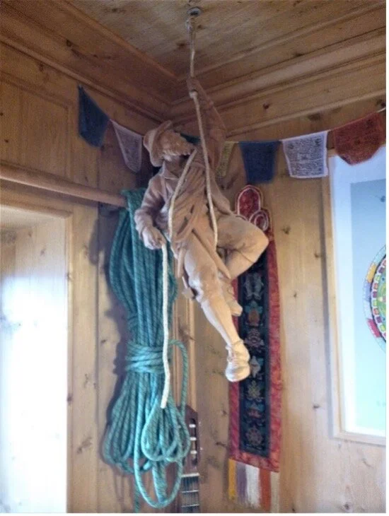 Eine Holzfigur eines Kletterers hängt an einem Seil von der Decke einer gemütlichen Berghütte. | © DAV Dortmund