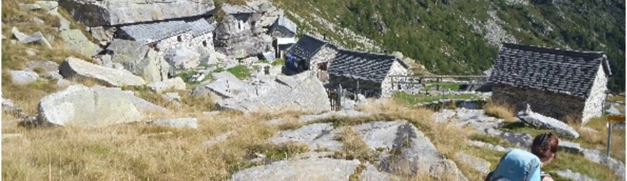 Wandernde Person durchquert ein abgelegenes Steindorf in einer alpinen Landschaft mit steilen Hängen. | © DAV Dortmund