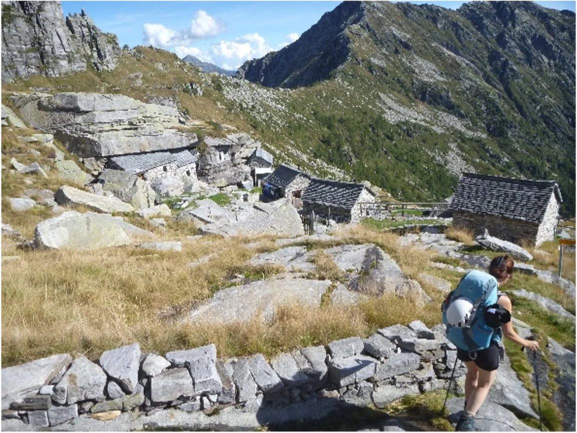 Wandernde Person durchquert ein abgelegenes Steindorf in einer alpinen Landschaft mit steilen Hängen. | © DAV Dortmund
