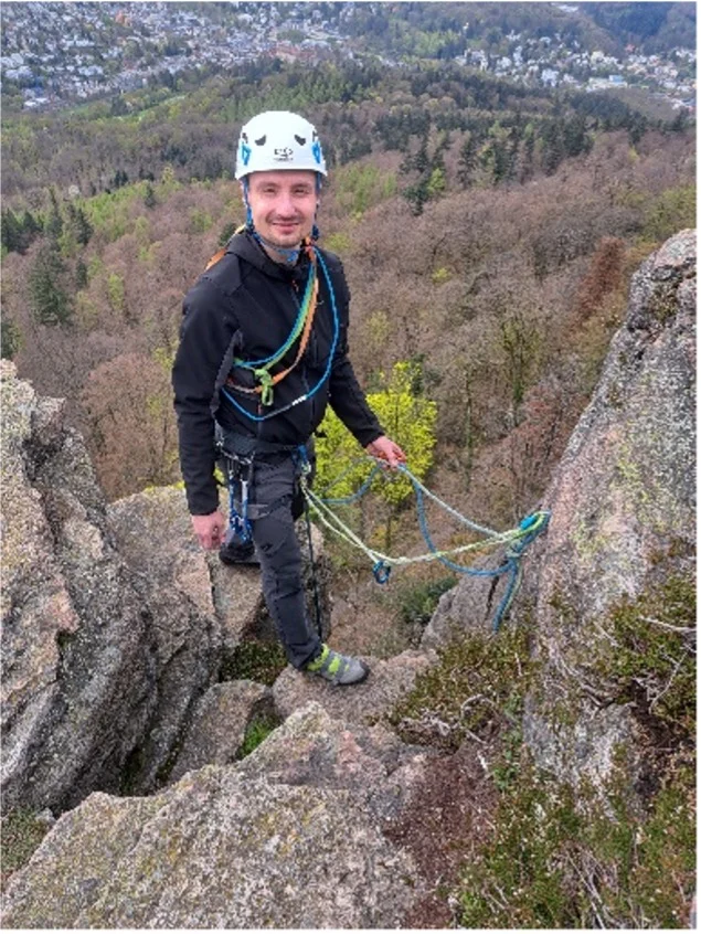 Eine Person mit Kletterhelm und Klettergurt steht auf einem Felsgrat mit Ausblick auf ein bewaldetes Tal. | © DAV Dortmund