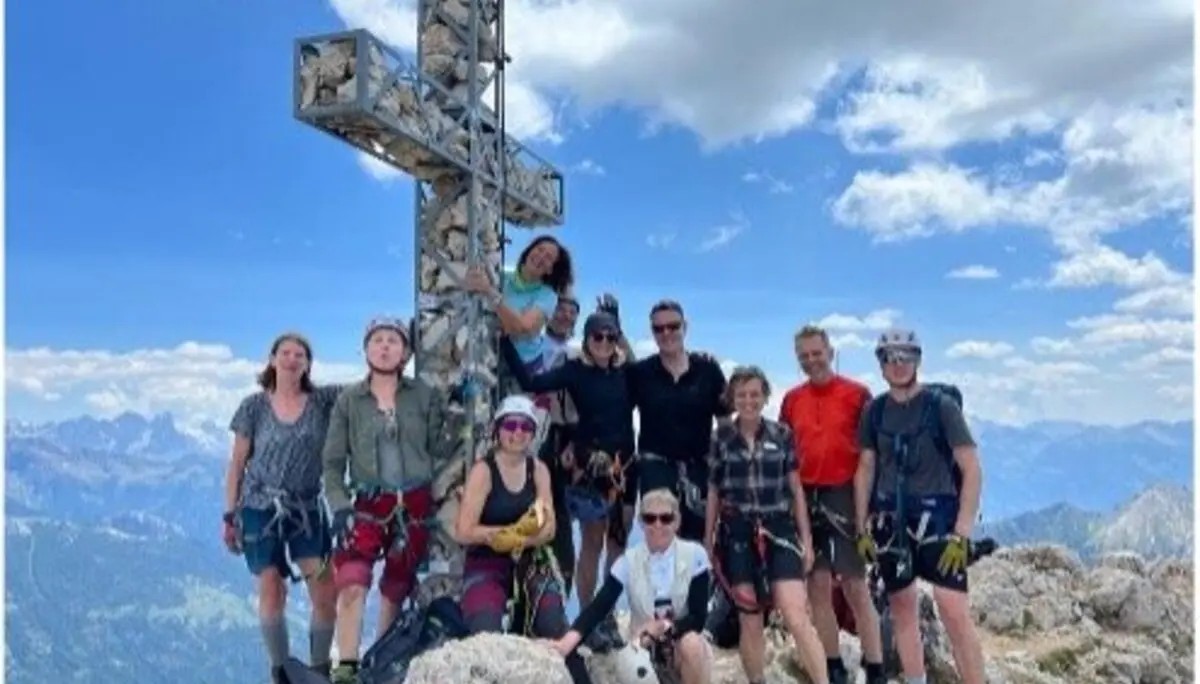  Alt-Text: Zehn Personen stehen lachend am Gipfelkreuz mit beeindruckendem Panorama und blauem Himmel. | © DAV Dortmund