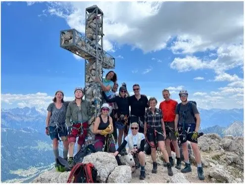  Alt-Text: Zehn Personen stehen lachend am Gipfelkreuz mit beeindruckendem Panorama und blauem Himmel. | © DAV Dortmund
