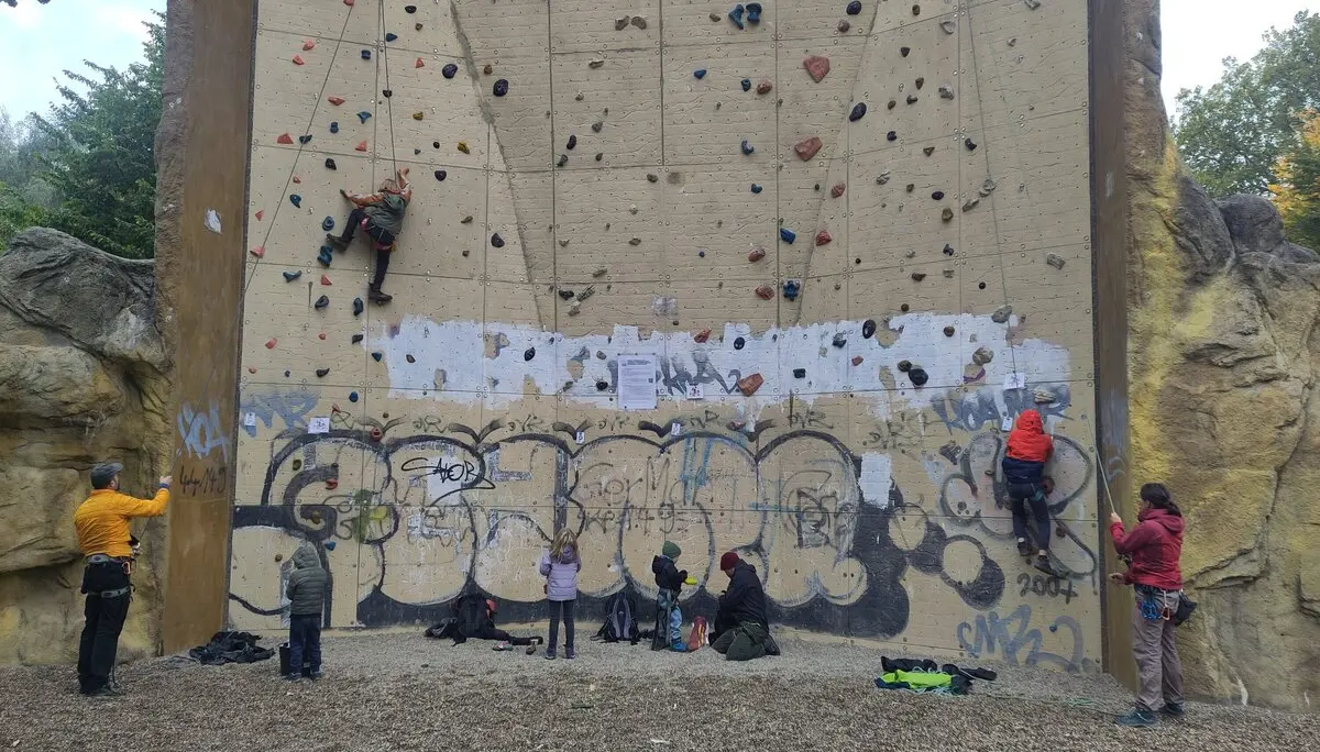 Mehrere Personen klettern und sichern am Kletterturm Dortmund. Kinder und Erwachsene üben gemeinsam an der künstlichen Kletterwand im Freien. Die Wand ist mit bunten Griffen ausgestattet und teilweise mit Graffiti versehen. | © DAV Dortmund