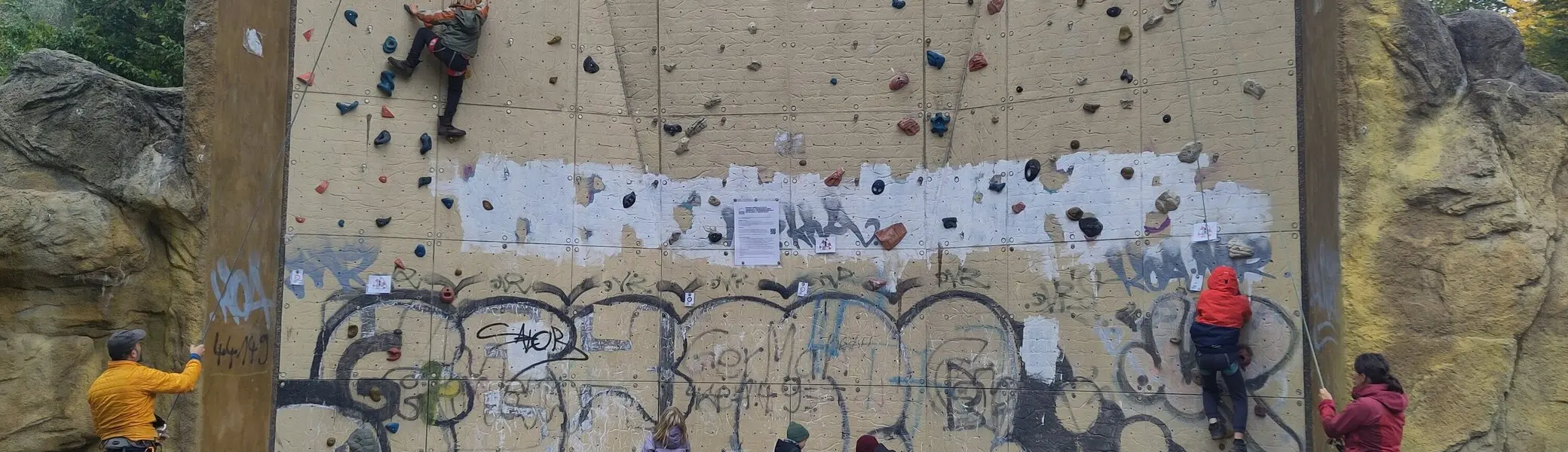 Mehrere Personen klettern und sichern am Kletterturm Dortmund. Kinder und Erwachsene üben gemeinsam an der künstlichen Kletterwand im Freien. Die Wand ist mit bunten Griffen ausgestattet und teilweise mit Graffiti versehen. | © DAV Dortmund