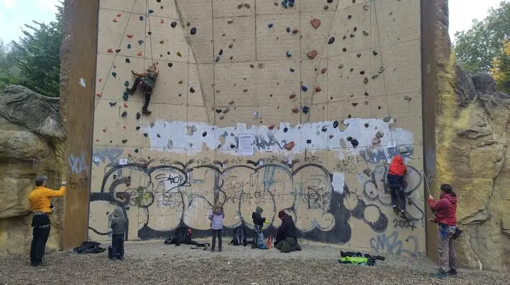 Mehrere Personen klettern und sichern am Kletterturm Dortmund. Kinder und Erwachsene üben gemeinsam an der künstlichen Kletterwand im Freien. Die Wand ist mit bunten Griffen ausgestattet und teilweise mit Graffiti versehen. | © DAV Dortmund