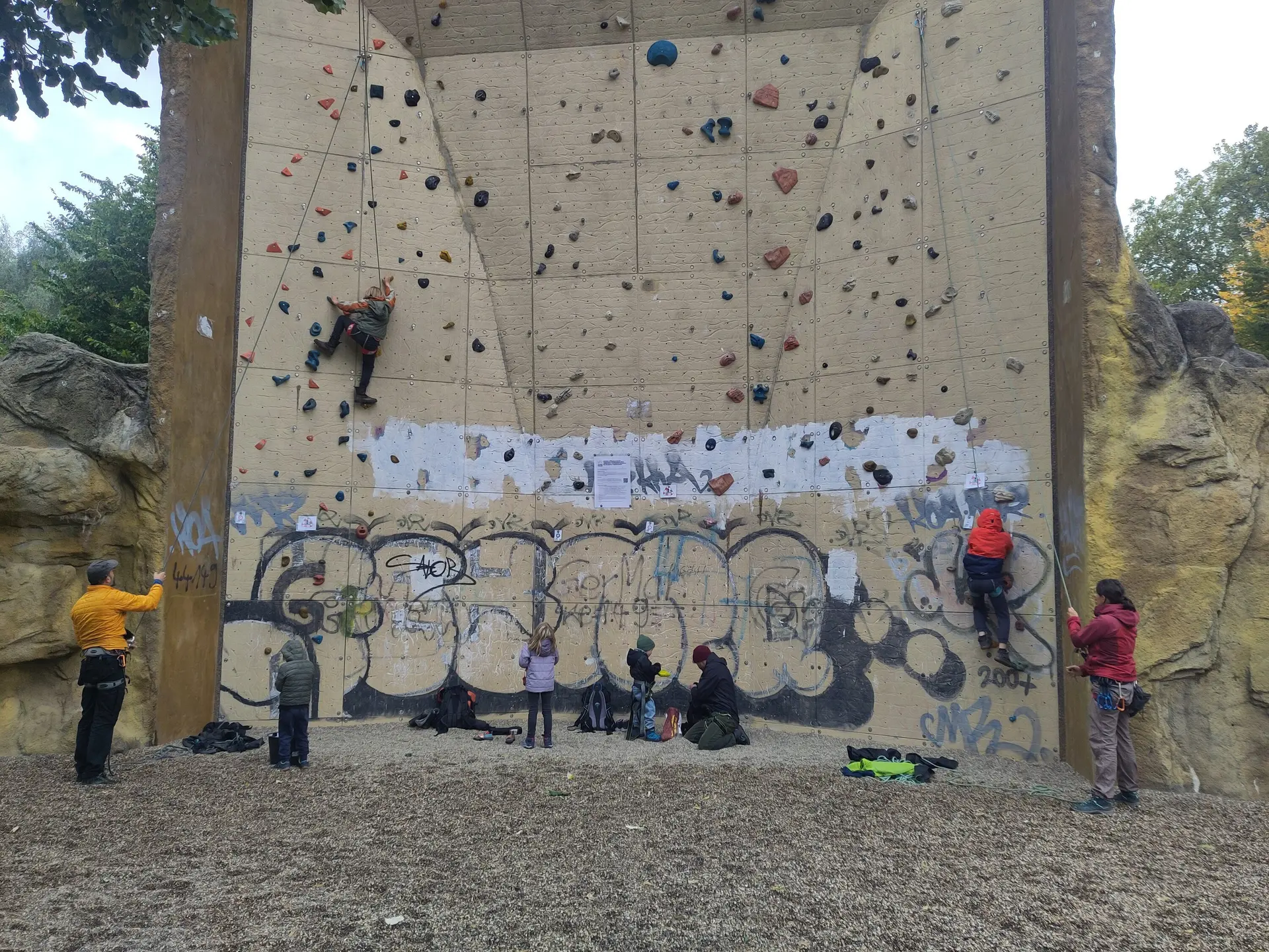 Mehrere Personen klettern und sichern am Kletterturm Dortmund. Kinder und Erwachsene üben gemeinsam an der künstlichen Kletterwand im Freien. Die Wand ist mit bunten Griffen ausgestattet und teilweise mit Graffiti versehen. | © DAV Dortmund