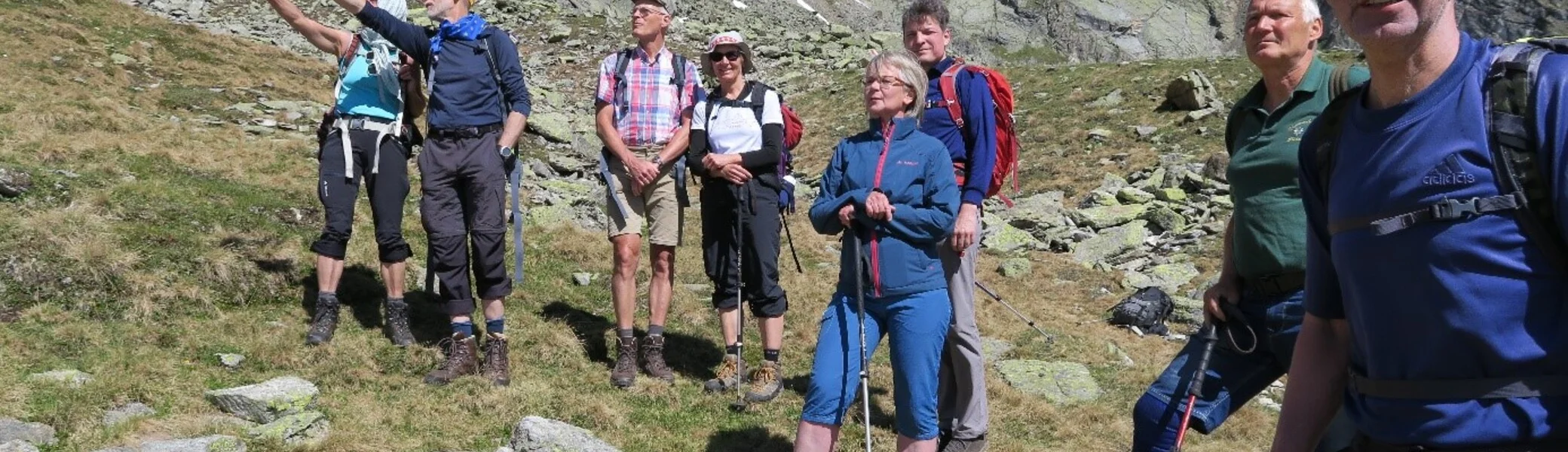Eine Gruppe steht auf einer Bergwiese, einige zeigen in Richtung Gipfel, andere schauen entspannt. | © DAV Dortmund