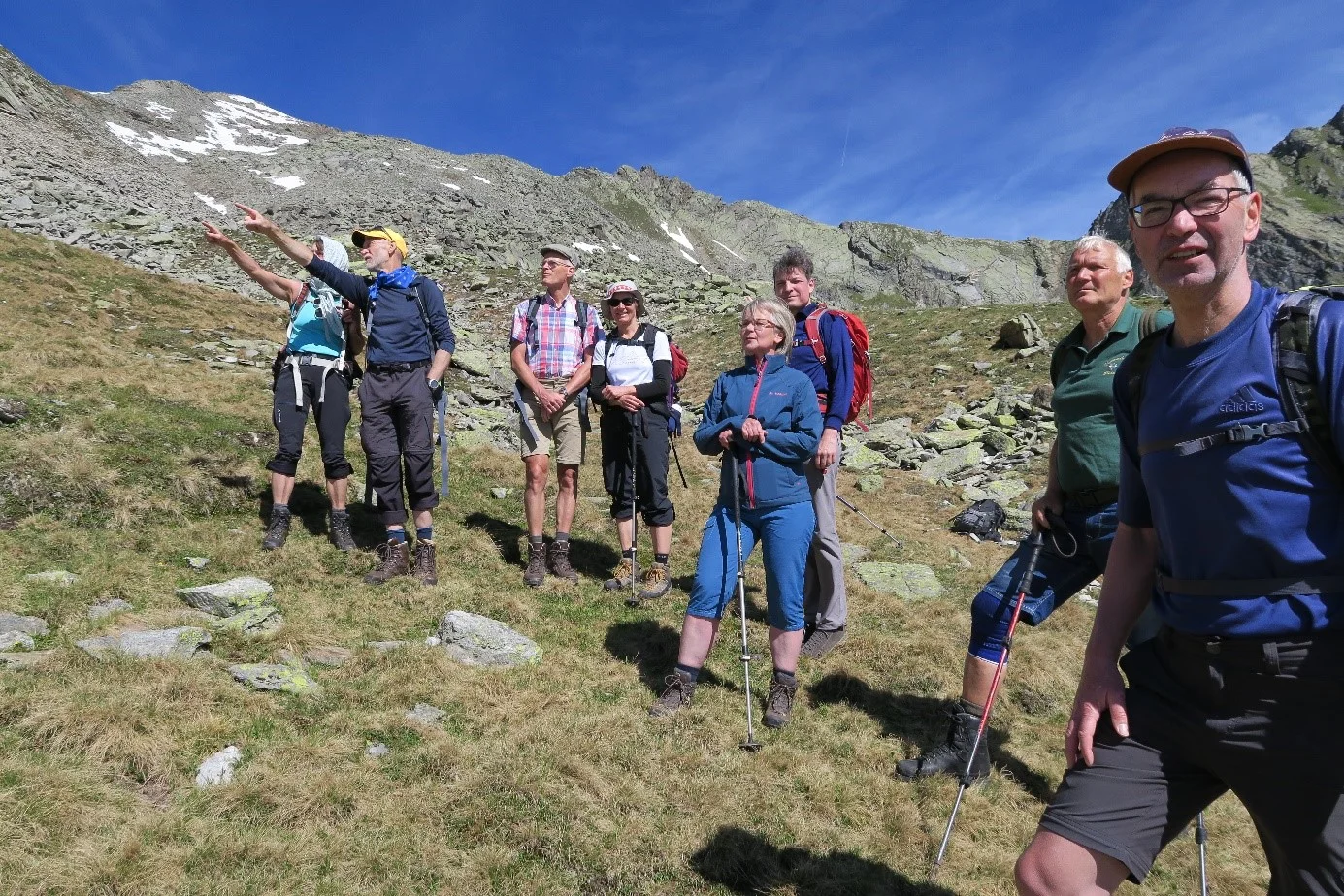 Eine Gruppe steht auf einer Bergwiese, einige zeigen in Richtung Gipfel, andere schauen entspannt. | © DAV Dortmund