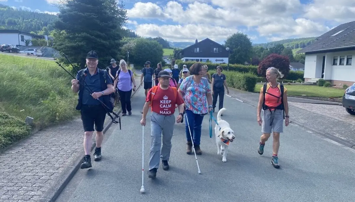 Eine gemischte Gruppe von Menschen mit und ohne Sehbehinderung wandert gemeinsam mit zwei Hunden auf einer Dorfstraße bei sonnigem Wetter. | © DAV Dortmund