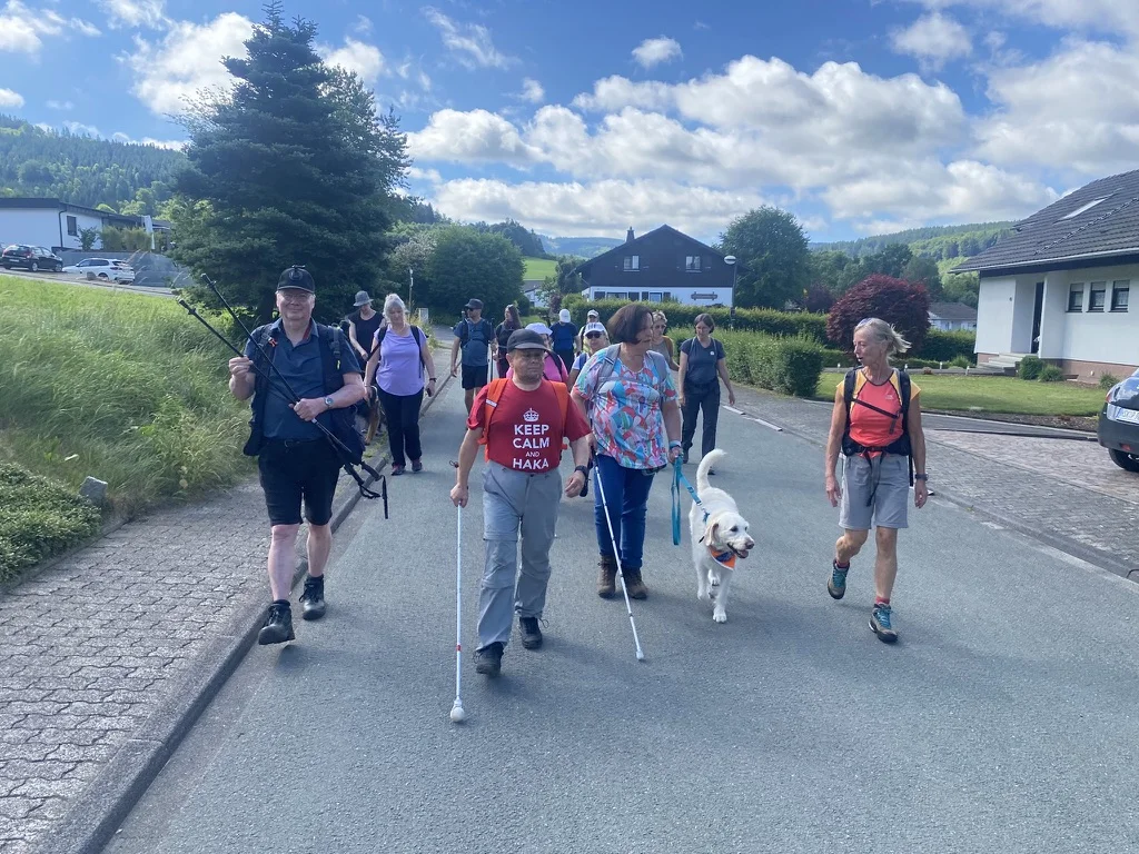 Eine gemischte Gruppe von Menschen mit und ohne Sehbehinderung wandert gemeinsam mit zwei Hunden auf einer Dorfstraße bei sonnigem Wetter. | © DAV Dortmund