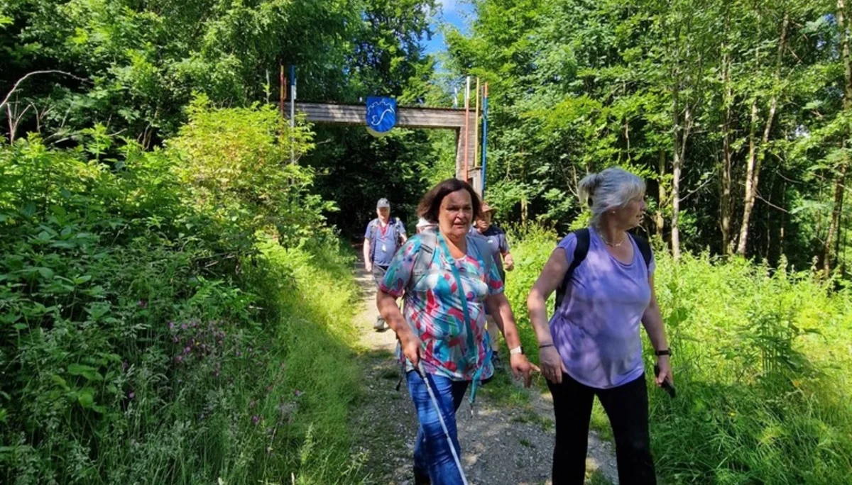 Zwei Frauen, eine davon mit Langstock, wandern auf einem schmalen Pfad im Wald. Im Hintergrund ist ein Holzbogen mit einem blauen Wappen sichtbar. | © DAV Dortmund