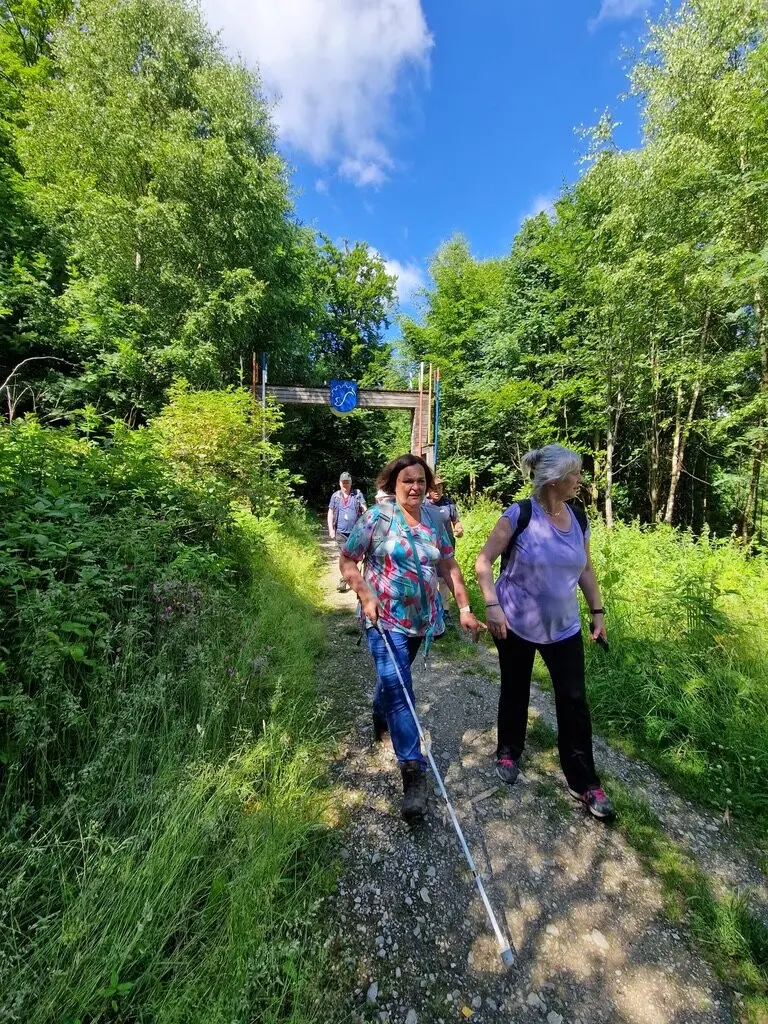 Zwei Frauen, eine davon mit Langstock, wandern auf einem schmalen Pfad im Wald. Im Hintergrund ist ein Holzbogen mit einem blauen Wappen sichtbar. | © DAV Dortmund