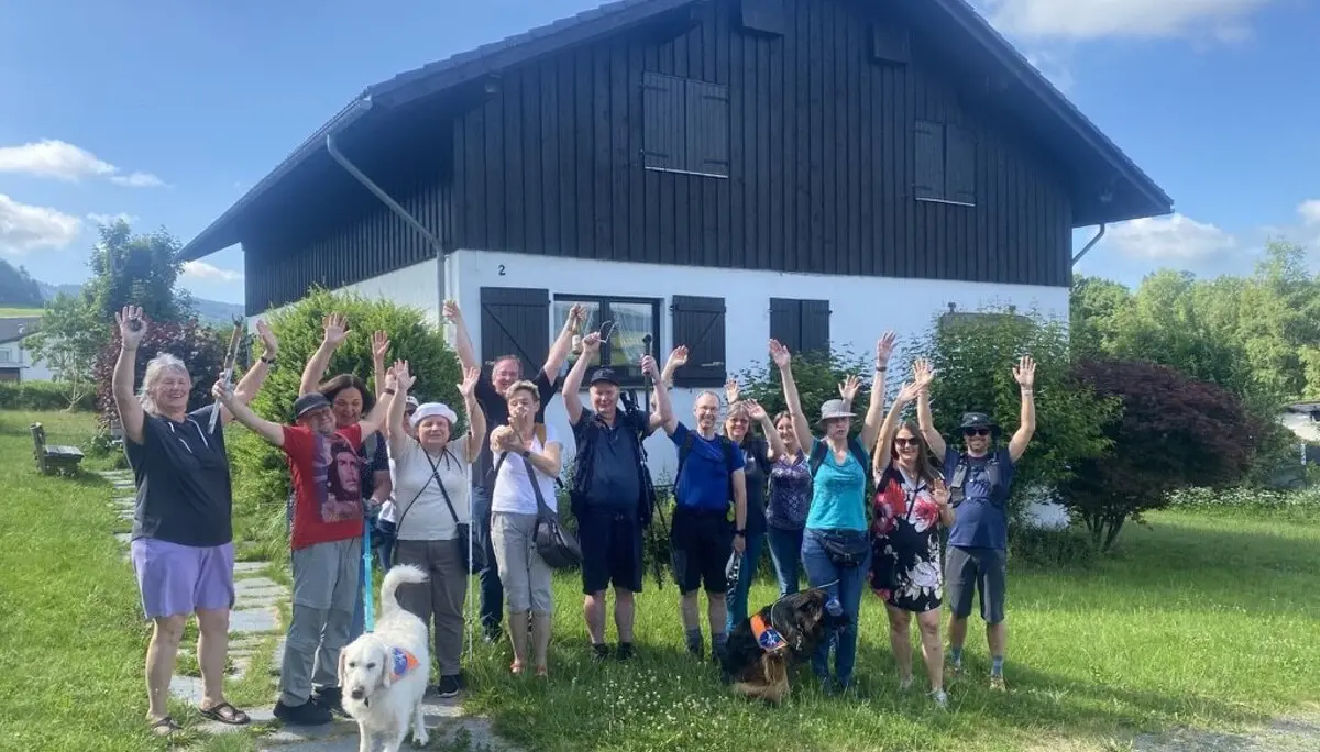 Die gesamte Wandergruppe steht vor einem schwarzen Holzhaus und winkt lachend in die Kamera. Zwei Hunde sind ebenfalls dabei. | © DAV Dortmund