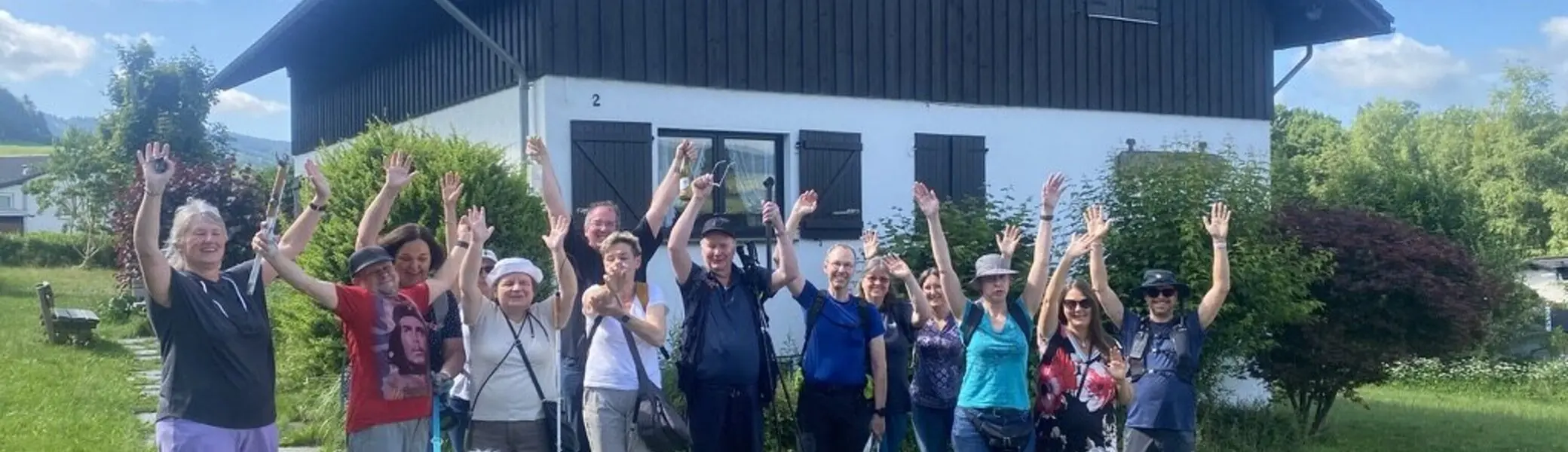 Die gesamte Wandergruppe steht vor einem schwarzen Holzhaus und winkt lachend in die Kamera. Zwei Hunde sind ebenfalls dabei. | © DAV Dortmund