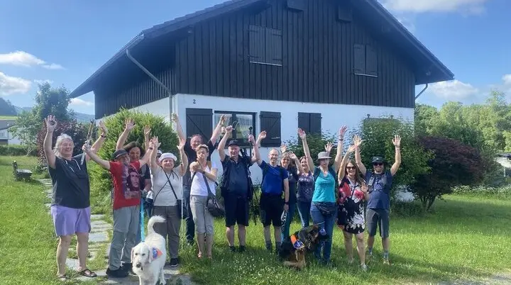 Die gesamte Wandergruppe steht vor einem schwarzen Holzhaus und winkt lachend in die Kamera. Zwei Hunde sind ebenfalls dabei. | © DAV Dortmund