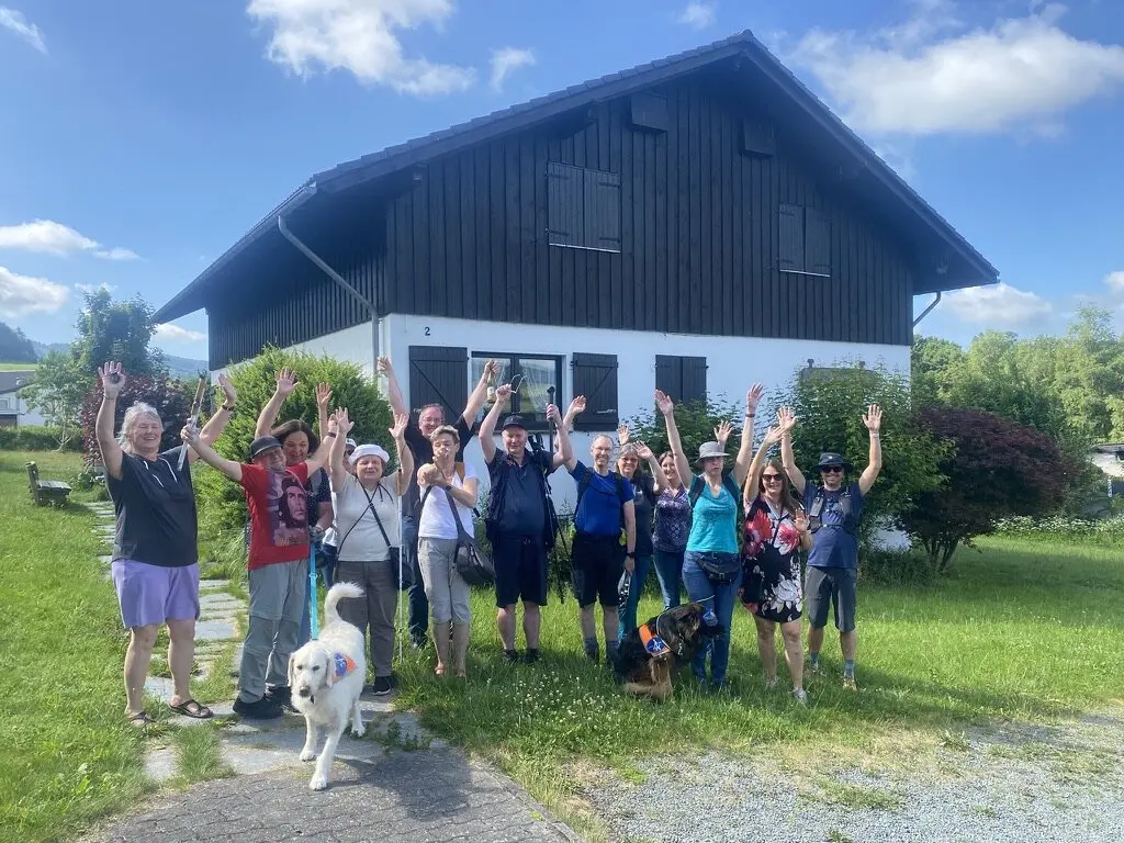 Die gesamte Wandergruppe steht vor einem schwarzen Holzhaus und winkt lachend in die Kamera. Zwei Hunde sind ebenfalls dabei. | © DAV Dortmund