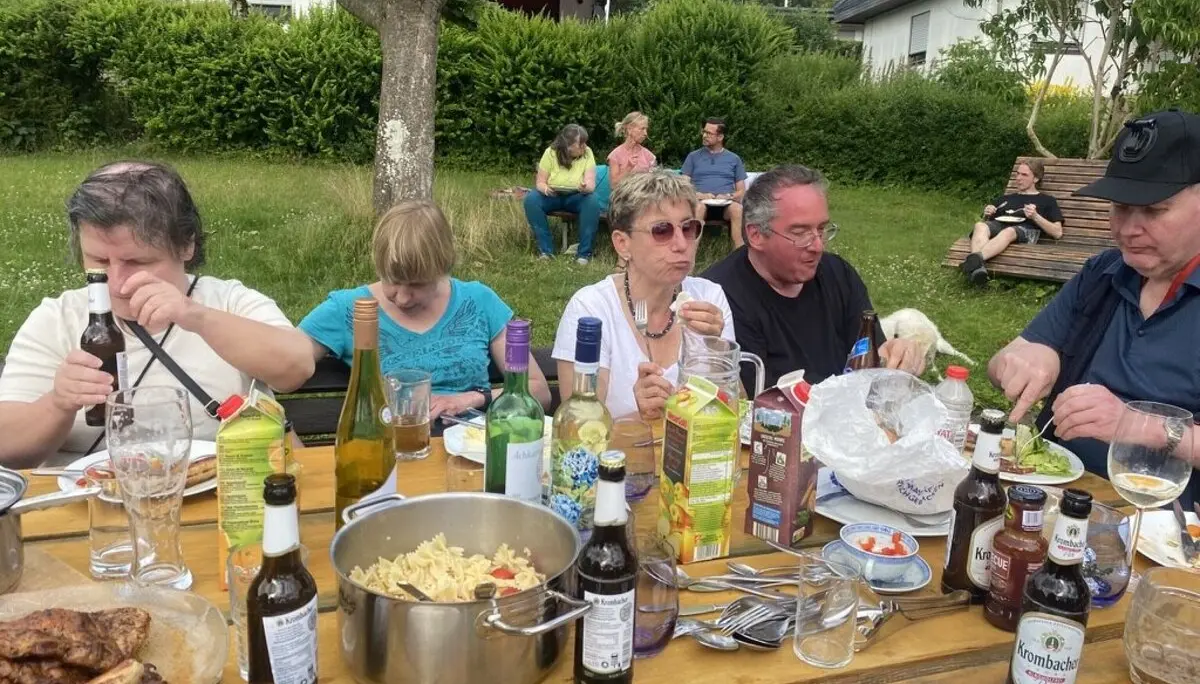 Mehrere Personen sitzen an einem Tisch im Garten und essen gemeinsam. Es gibt Würstchen, Nudelsalat, Getränke und verschiedene Soßen. | © DAV Dortmund