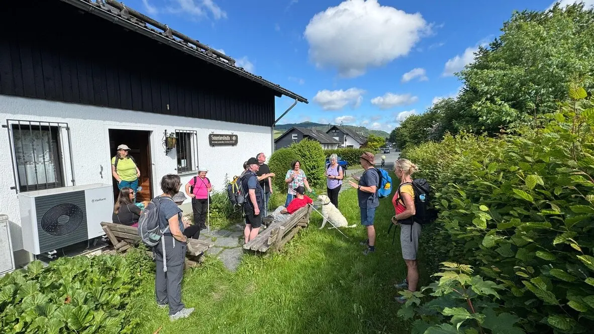 Die Gruppe steht bei Sonnenschein vor einem weißen Gebäude mit dem Schild „Sauerlandhütte“, ein blinder Teilnehmer sitzt mit Hund auf einer Bank. | © DAV Dortmund