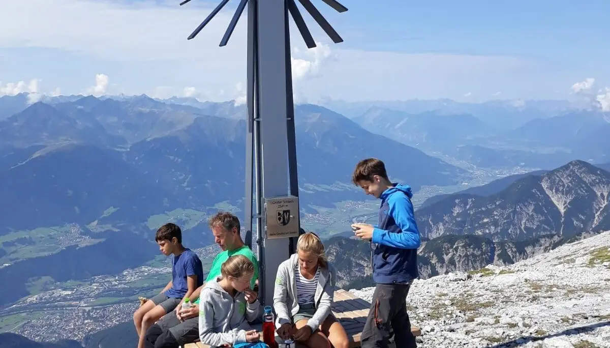 Fünf Jugendliche sitzen oder stehen entspannt an einem modernen Gipfelkreuz aus Metall mit Panoramablick ins Tal. | © DAV Dortmund