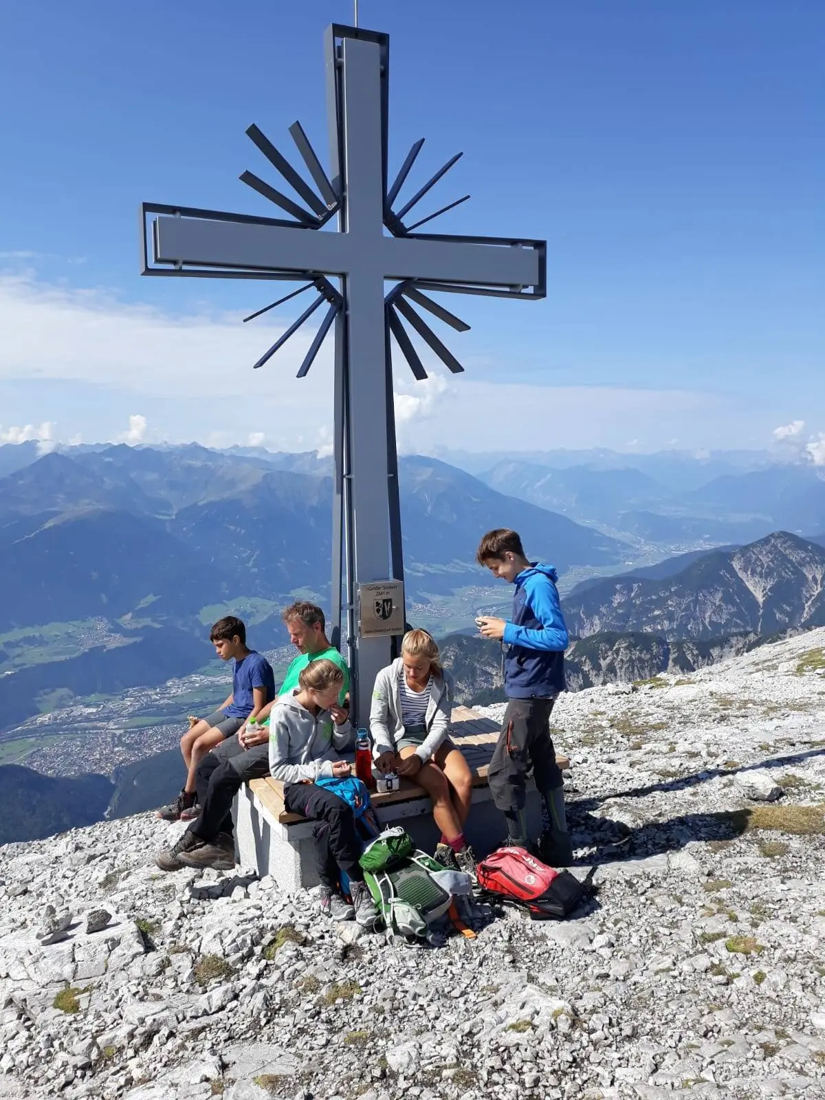 Fünf Jugendliche sitzen oder stehen entspannt an einem modernen Gipfelkreuz aus Metall mit Panoramablick ins Tal. | © DAV Dortmund