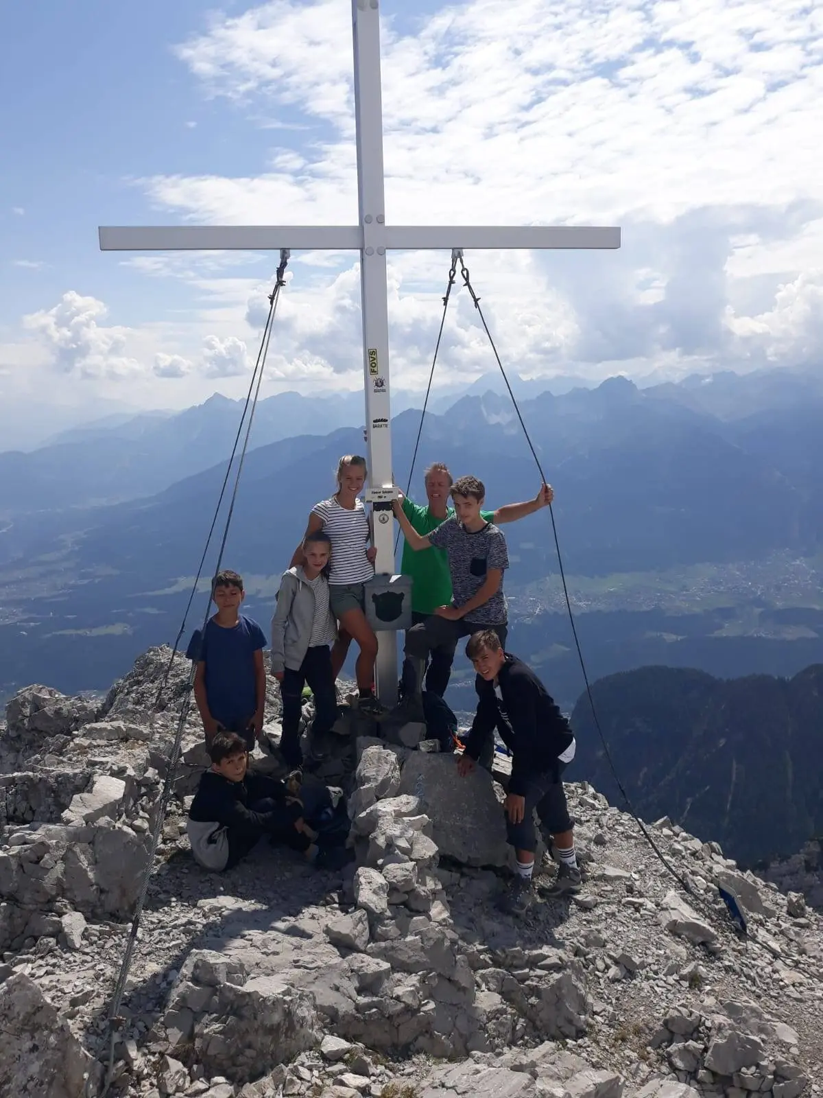 Mehrere Jugendliche stehen unter einem weißen Gipfelkreuz auf einem felsigen Berg mit weitem Talblick im Hintergrund. | © DAV Dortmund