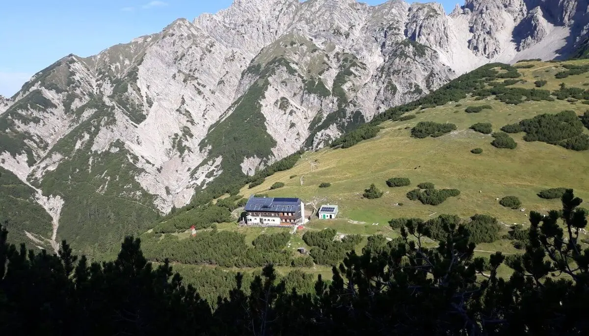 Eine Berghütte liegt auf einer grünen Wiese vor felsigem Bergmassiv unter blauem Himmel. | © DAV Dortmund