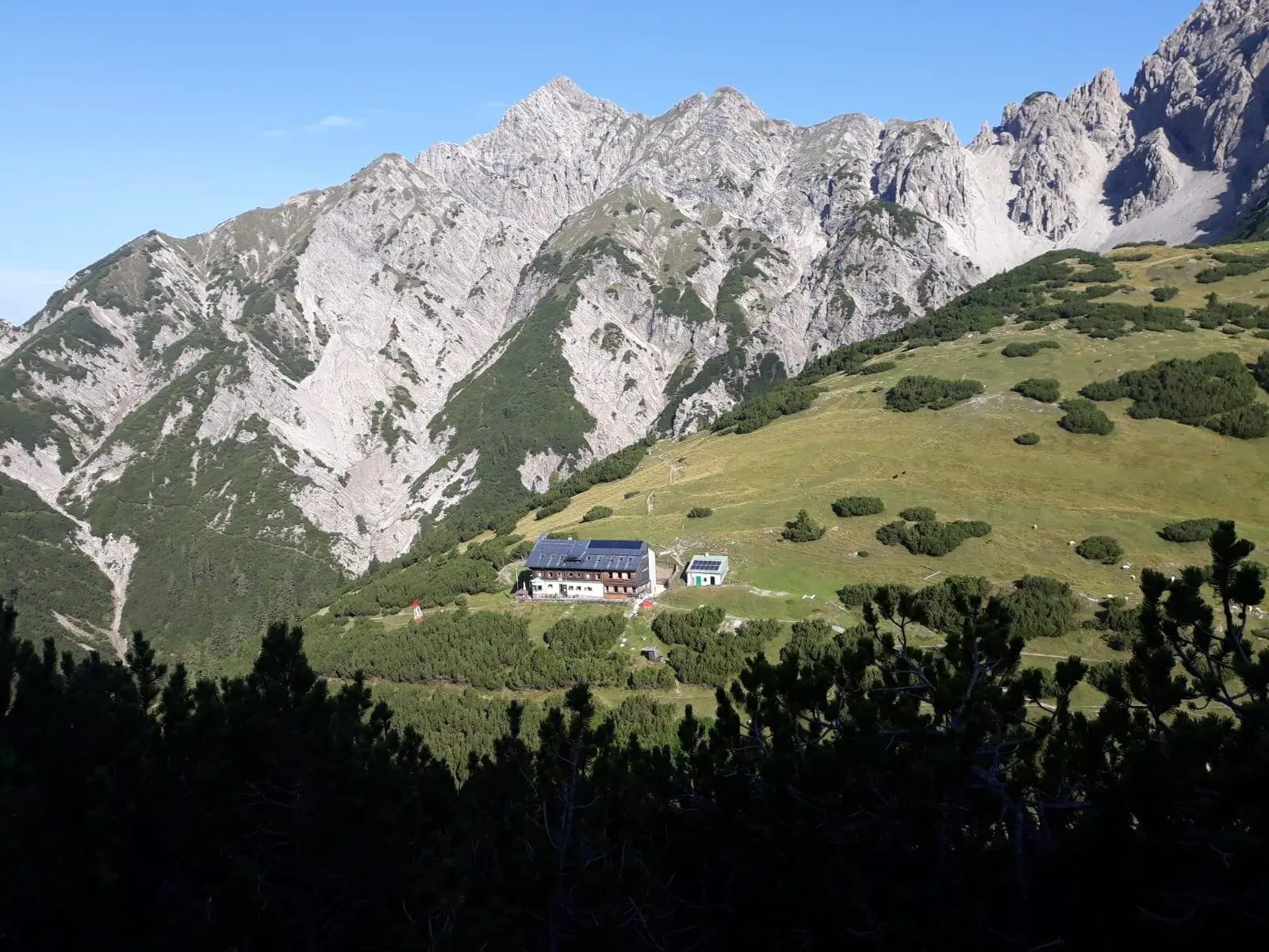 Eine Berghütte liegt auf einer grünen Wiese vor felsigem Bergmassiv unter blauem Himmel. | © DAV Dortmund