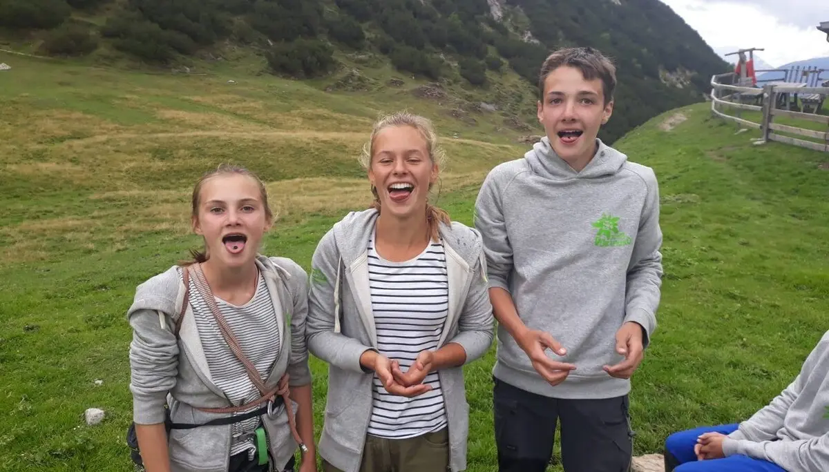 Drei Jugendliche stehen auf einer Almwiese, lachen und strecken die Zungen raus, im Hintergrund Berglandschaft. | © DAV Dortmund