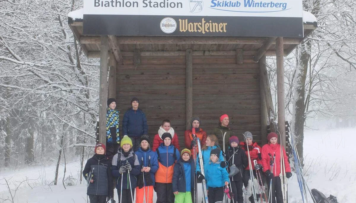 Gruppe von Jugendlichen mit Skiausrüstung vor einem hölzernen Unterstand mit Beschriftung "Biathlon Stadion Skiklub Winterberg". | © DAV Dortmund