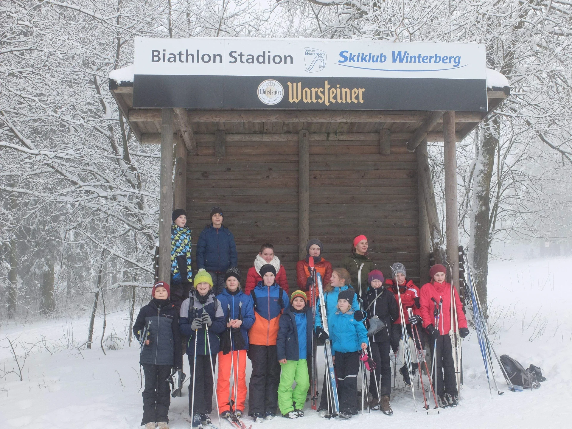 Gruppe von Jugendlichen mit Skiausrüstung vor einem hölzernen Unterstand mit Beschriftung "Biathlon Stadion Skiklub Winterberg". | © DAV Dortmund