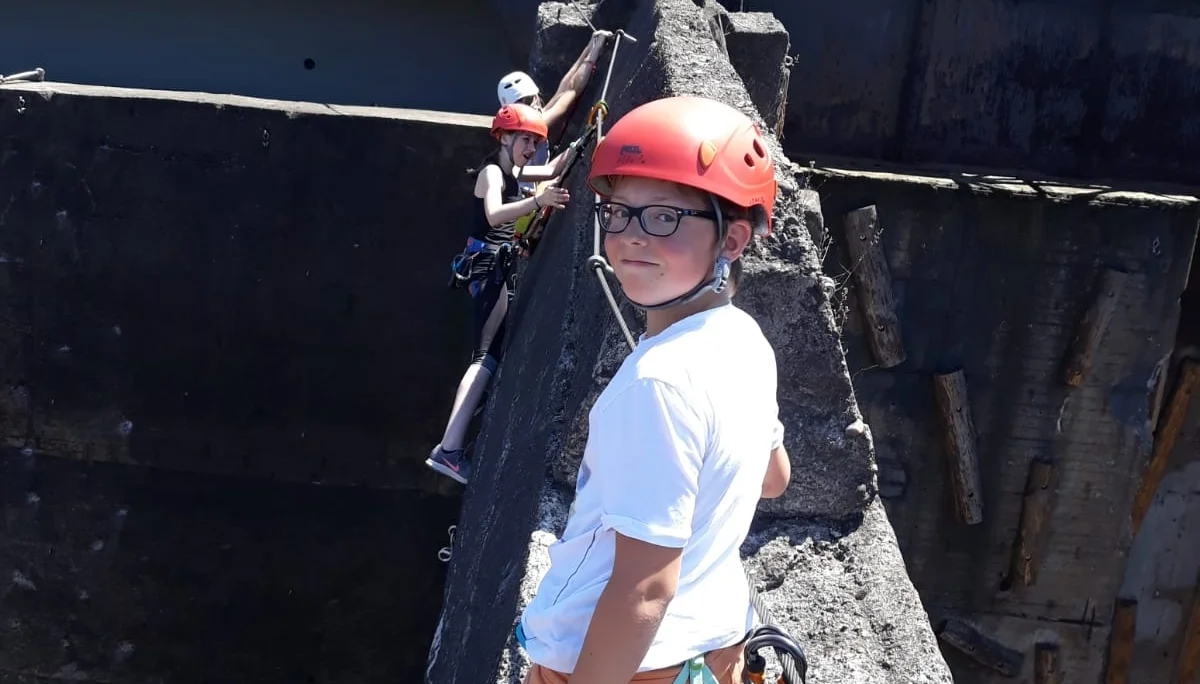 Zwei Kinder mit Helm und Klettergurt stehen gesichert auf einem Betonsteg zwischen alten Industrieanlagen. | © DAV Dortmund